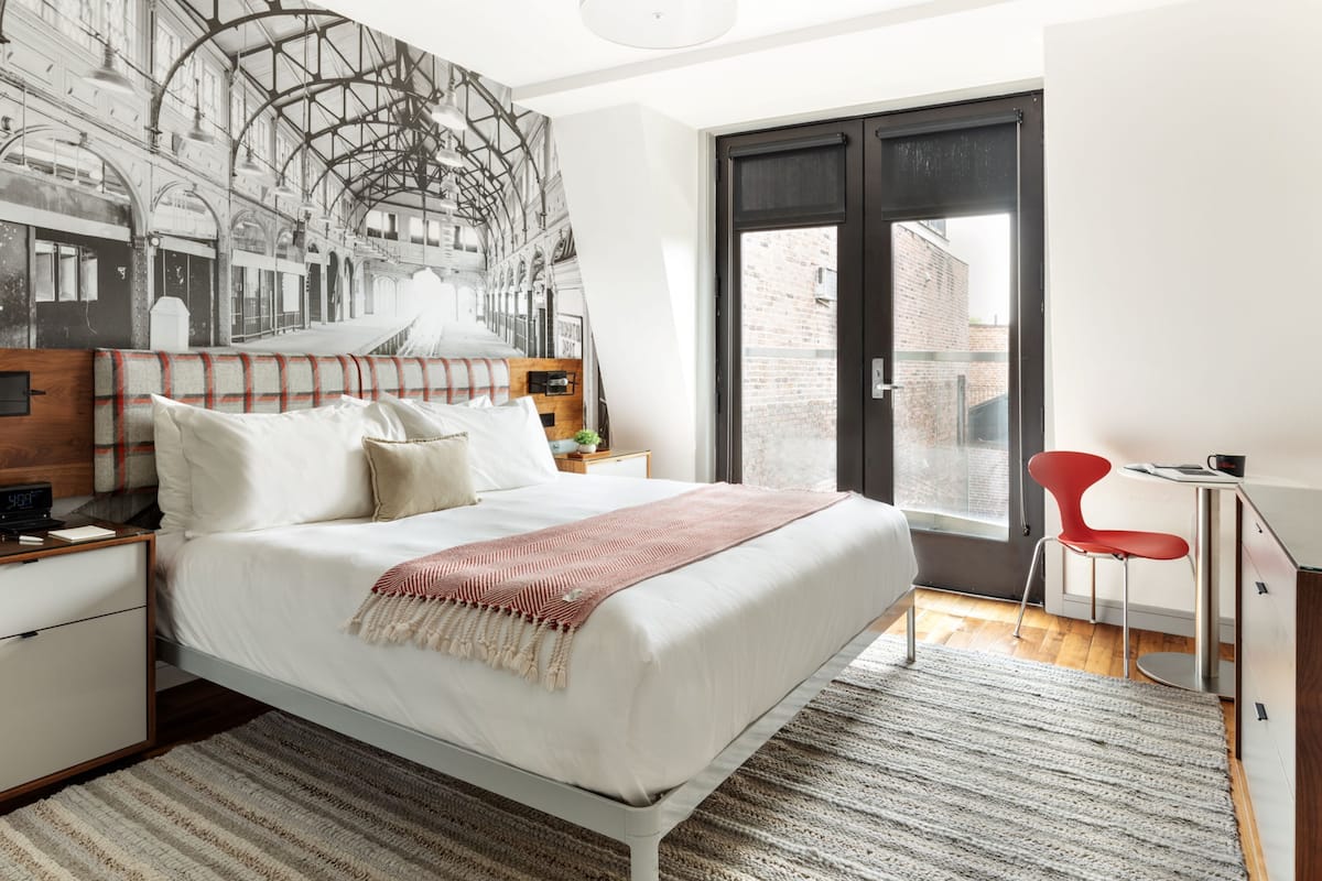 Ein modernes Schlafzimmer mit einem großen Bett, einem roten Sessel, einem Schreibtisch und einem Wandgemälde mit Stadtansicht.