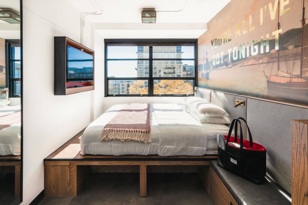 Ein modernes Schlafzimmer mit großem Fenster, einem Bett mit weißer Bettwäsche, einem an der Wand montierten Fernseher und einem Poster.