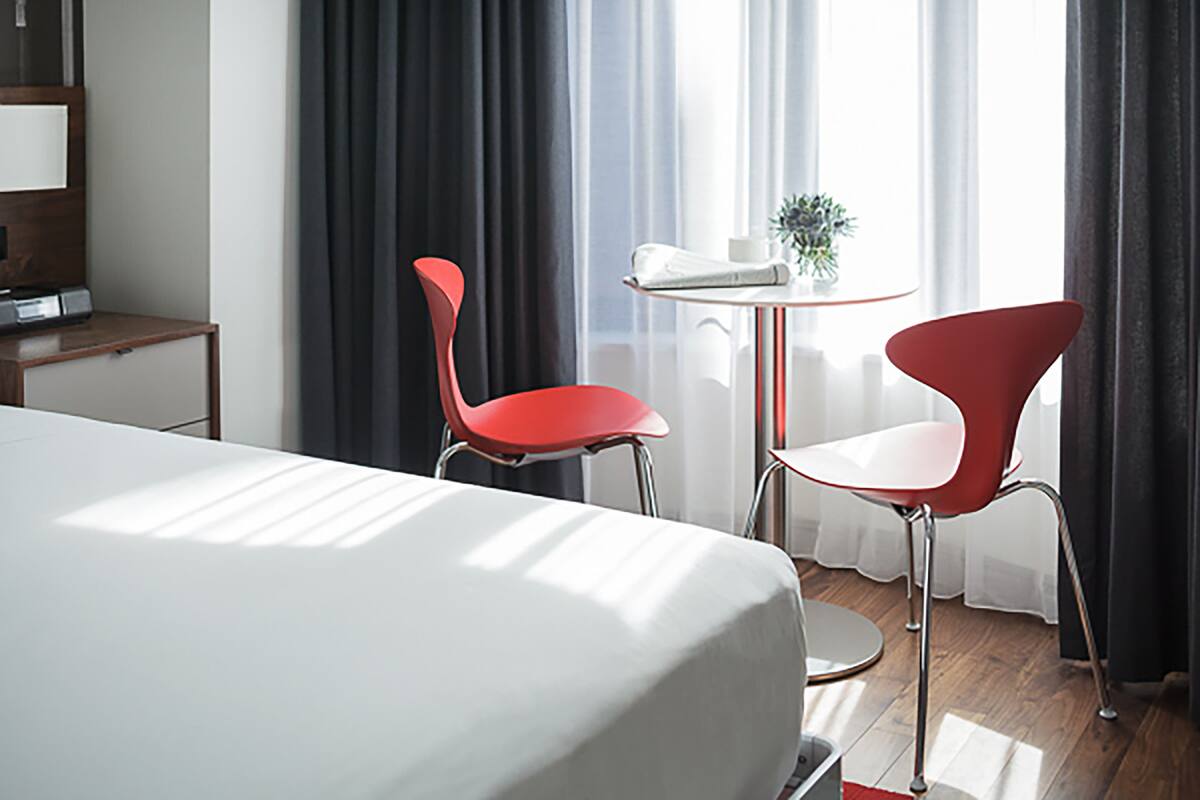 Ein Hotelzimmer mit einem Bett, zwei roten Sesseln, einem kleinen Tisch mit einer Pflanze und einem Fenster mit Vorhängen.