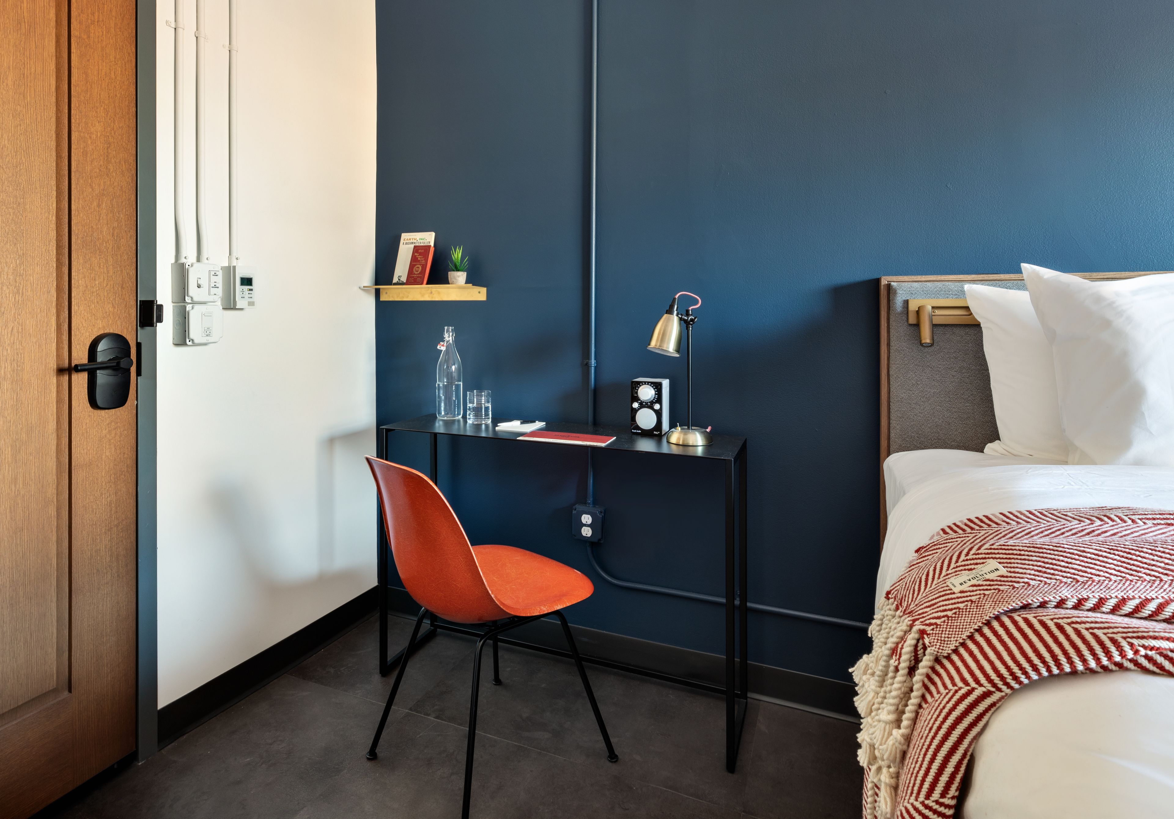A hotel room with a wooden door, a desk with a chair, a bed with white bedding, a red and white striped blanket, a small shelf with books and a plant, and a wall-mounted clock.