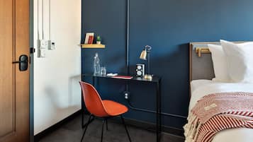 A hotel room with a wooden door, a desk with a chair, a bed with white bedding, a red and white striped blanket, a small shelf with books and a plant, and a wall-mounted clock.