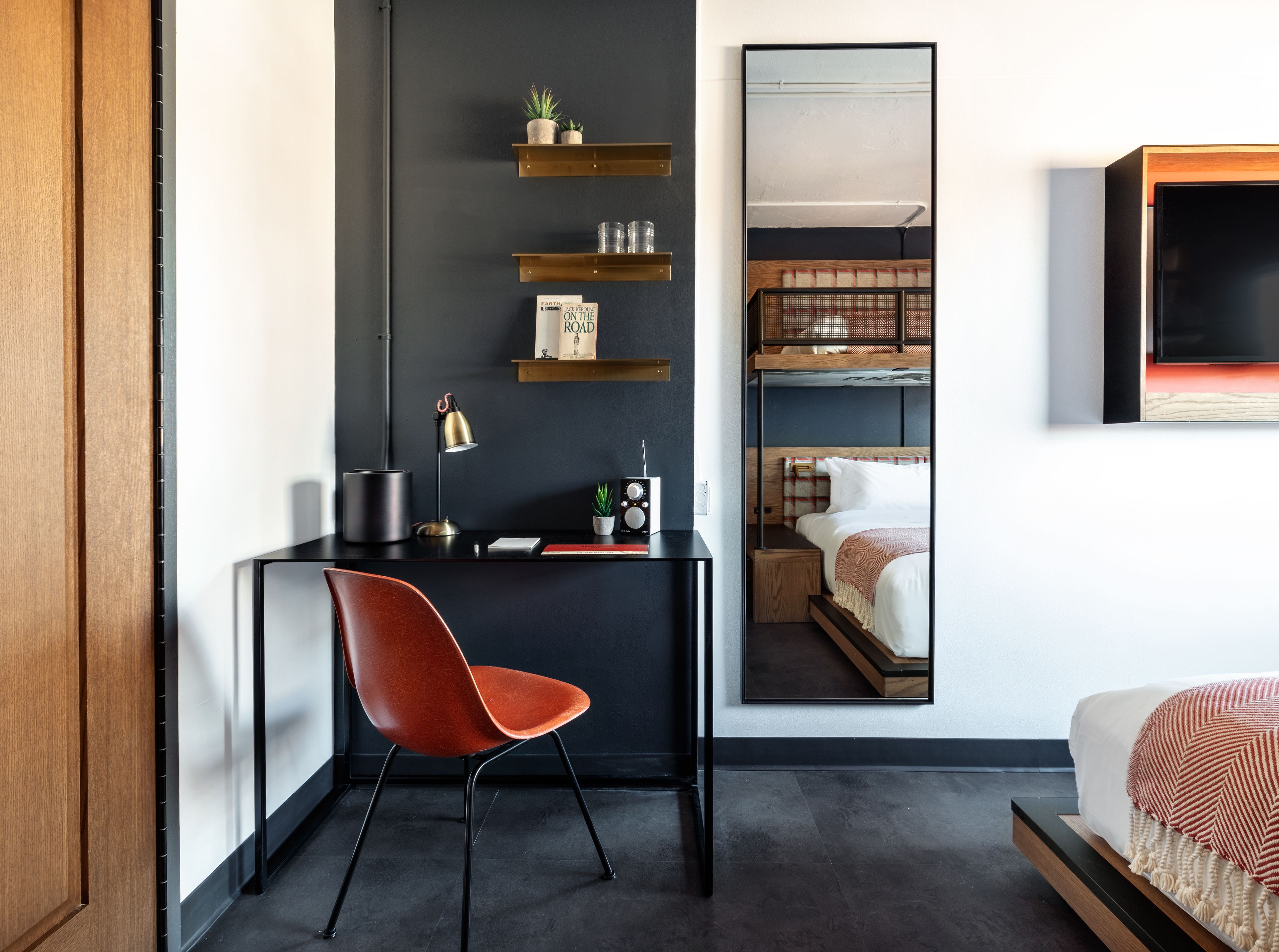 A modern hotel room with a dark wall, a wooden desk, a red chair, a bunk bed, and a mounted TV.