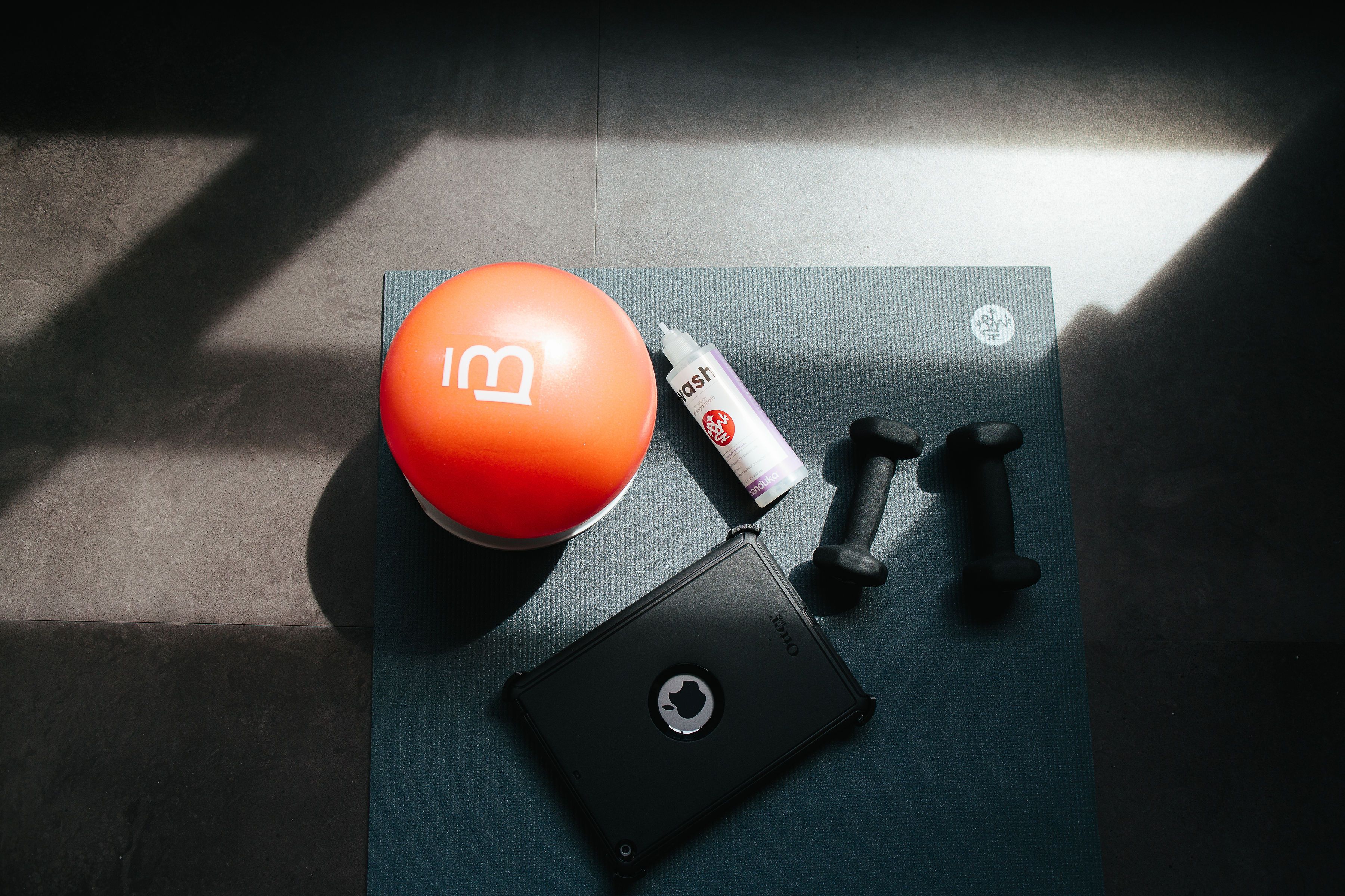 Una laptop negra con un logo circular, una botella roja y blanca, dos mancuernas negras y una colchoneta verde sobre una superficie oscura.