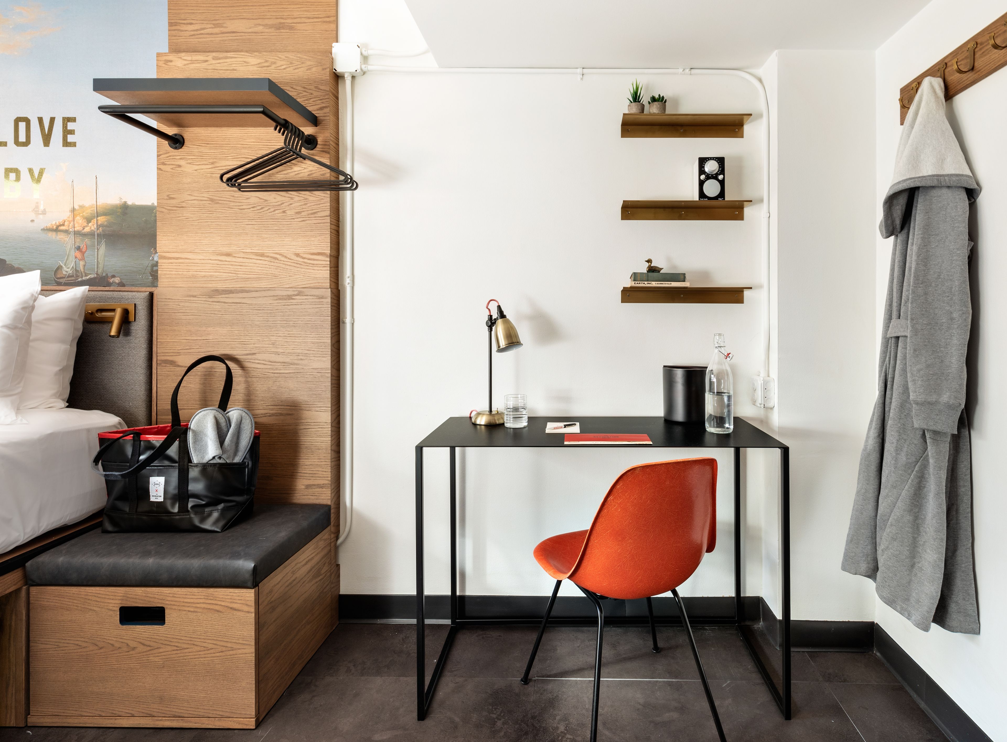 A modern hotel room with a wooden bed, a desk with a chair, a small shelf with plants, and a towel hanging on the wall.