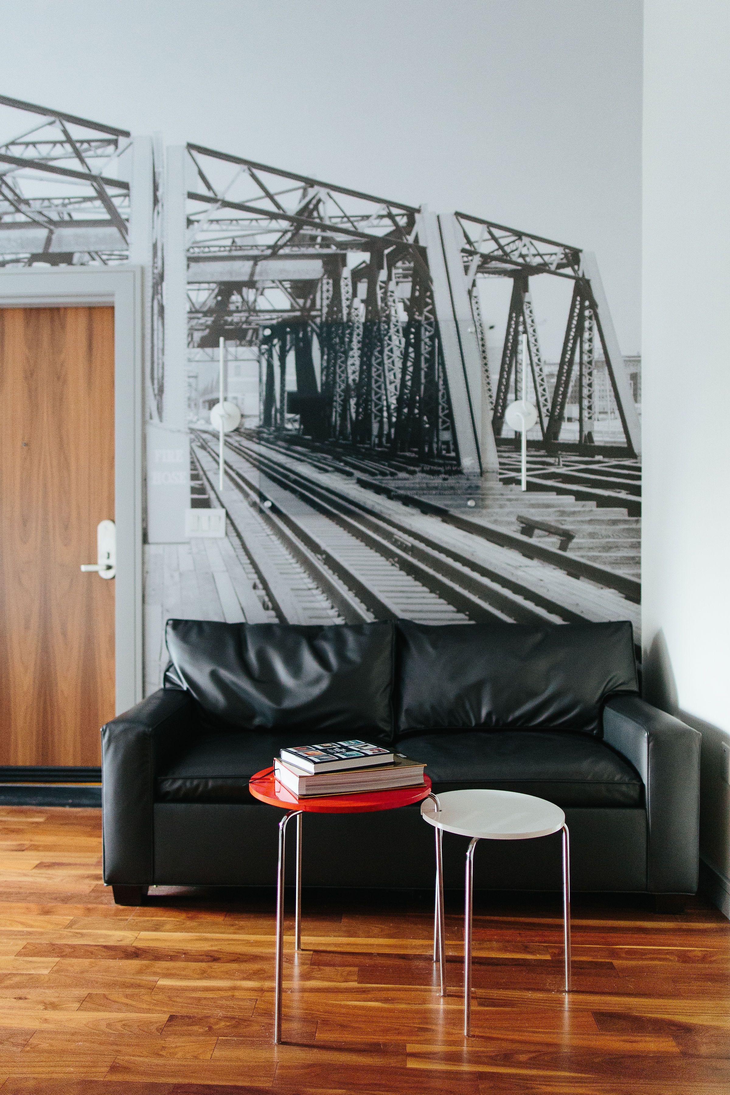 A modern living room with a black leather sofa, a small round table with a red base, and a large black and white photograph of a bridge on the wall.