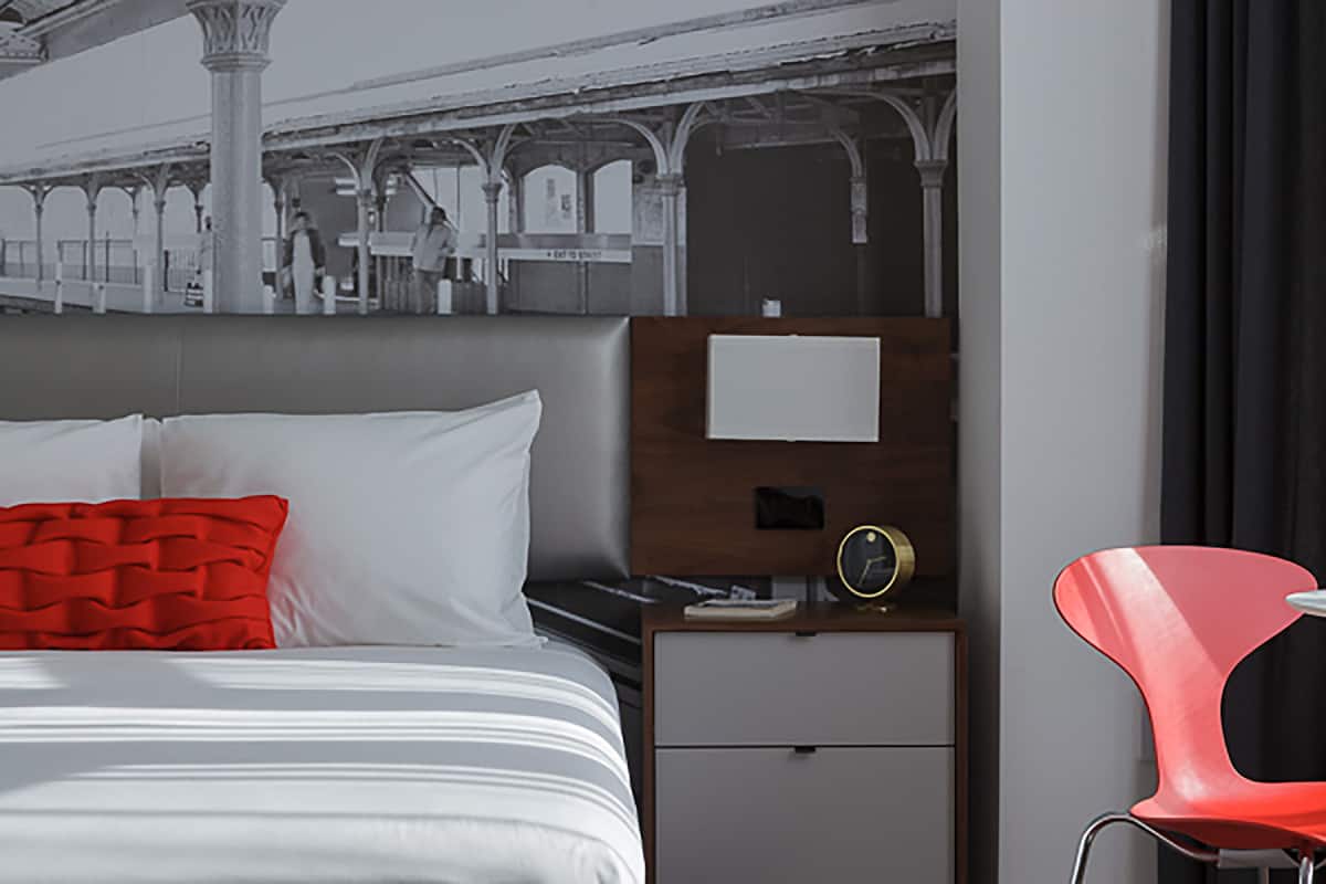 Ein Hotelzimmer mit einem Bett, einem Nachttisch, einem roten Sessel und einer Wanduhr.