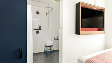 Egy modern fürőszoba zuhanyzóval, WC-vel és mosdókagylóval. Van benne egy kis szék és egy törölközőtartó.
