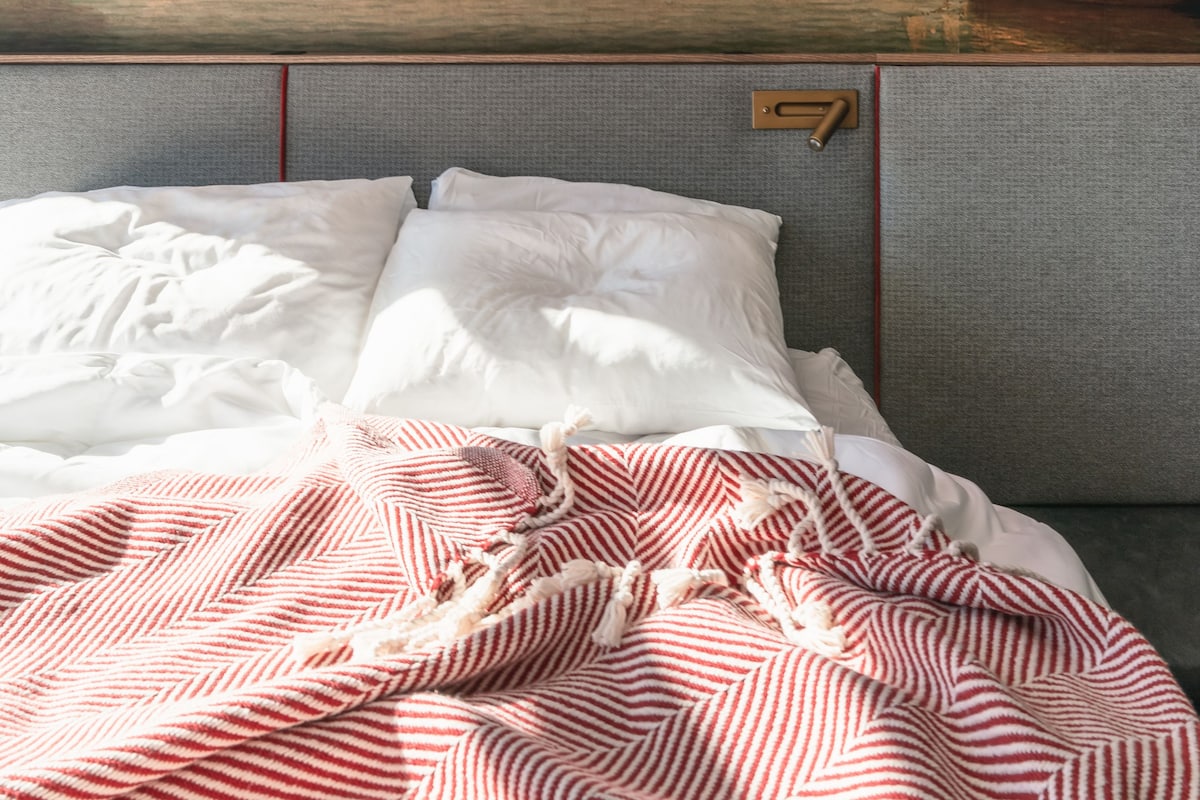 Ein Bett mit weißer Bettwäsche und einer rot-weiß gestreiften Decke, ein Kopfteil mit einer Holzvertäfelung und ein Wandbild mit einem Segelschiff auf dem Wasser.