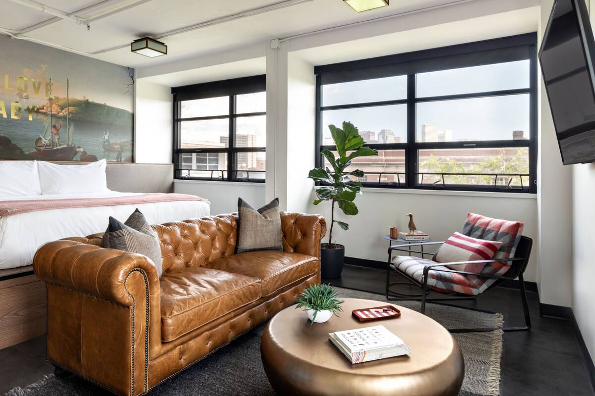 Ein modernes Hotelzimmer mit einem großen Bett, einem Ledersofa, einem kleinen runden Couchtisch und Blick auf die Stadt durch große Fenster.