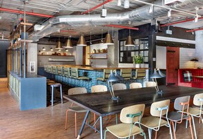 A modern restaurant with a long wooden table, multiple chairs, pendant lighting, and a bar area with stools.