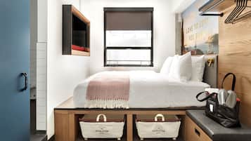 A compact hotel room with a bed, a window with a view, a wall-mounted TV, and a small seating area with a bag.