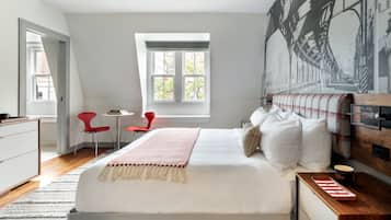 A modern bedroom with a large bed, a small table, two red chairs, and a window with a view of trees.