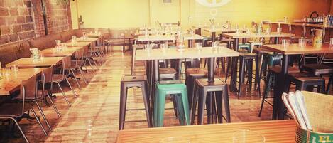 A spacious restaurant with multiple tables and chairs, a brick wall, and hanging plants.