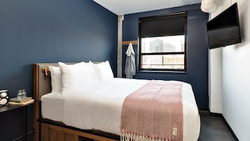 A bedroom with a bed, a nightstand, a suitcase, a window with a city view, and a TV on the wall.