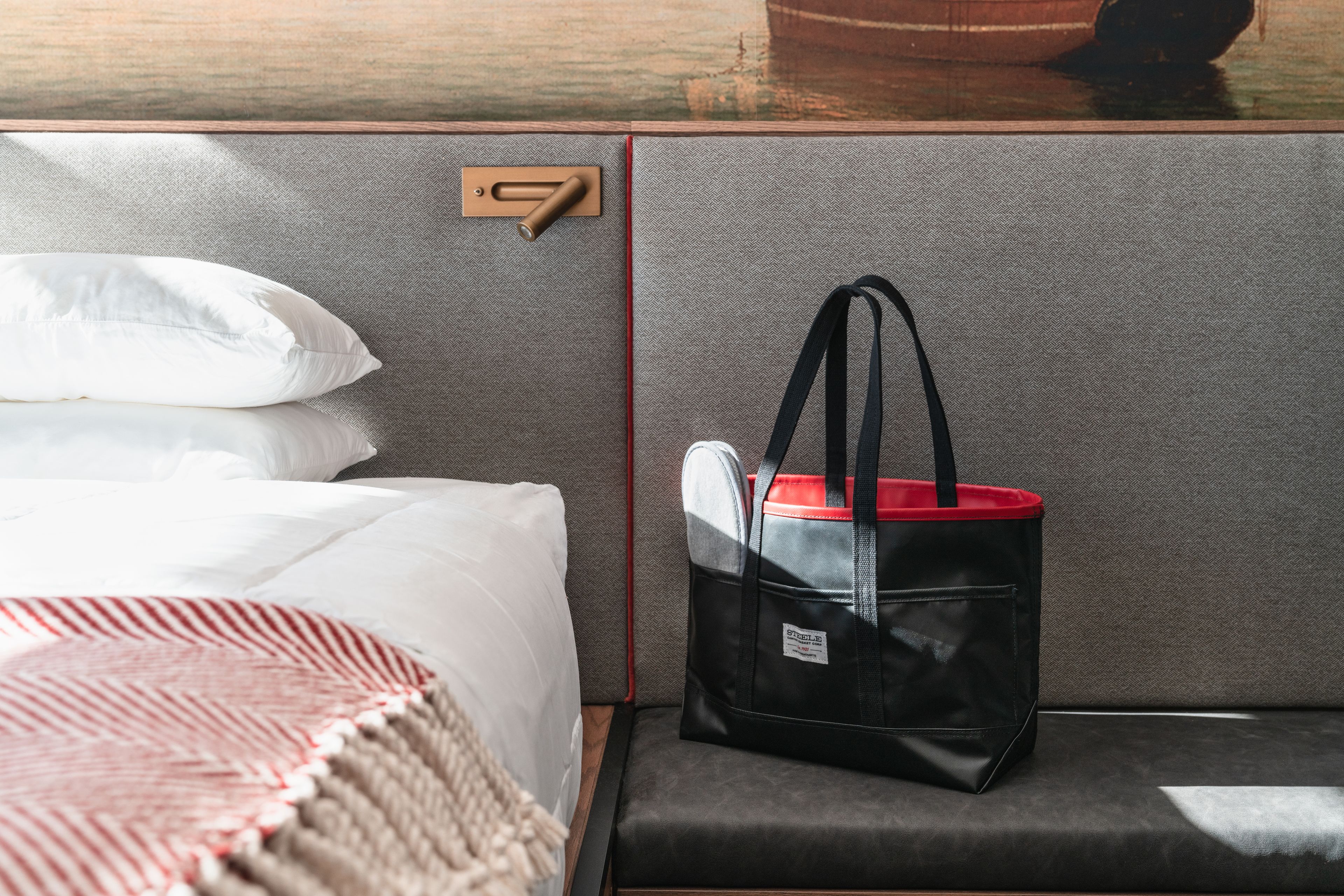 A hotel room with a bed, a grey upholstered bench, and a black tote bag with a red handle.