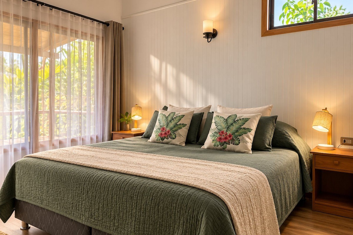 A bedroom with a bed, pillows, a nightstand, a lamp, a window with curtains, and a wall-mounted light fixture.