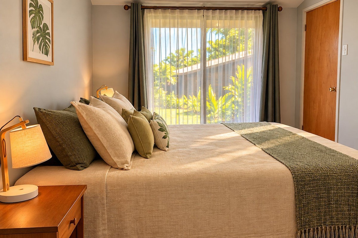 A bedroom with a bed, pillows, a bedside table with a lamp, a window with curtains, and a door.