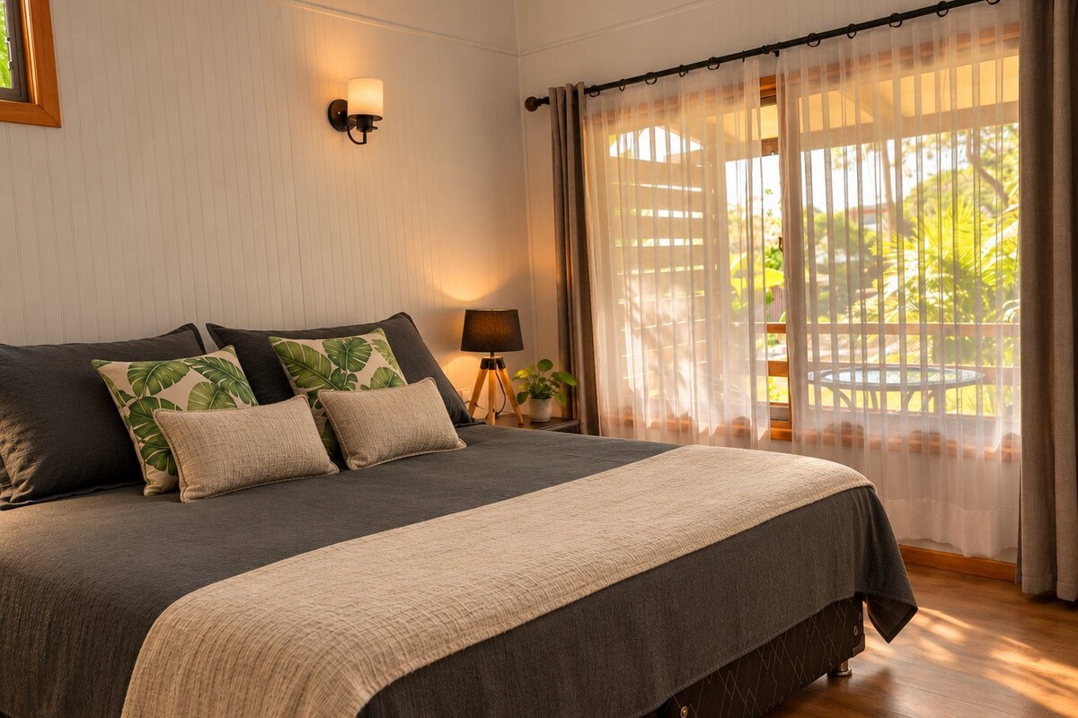 A bedroom with a large bed, a bedside lamp, a window with sheer curtains, and a view of greenery outside.