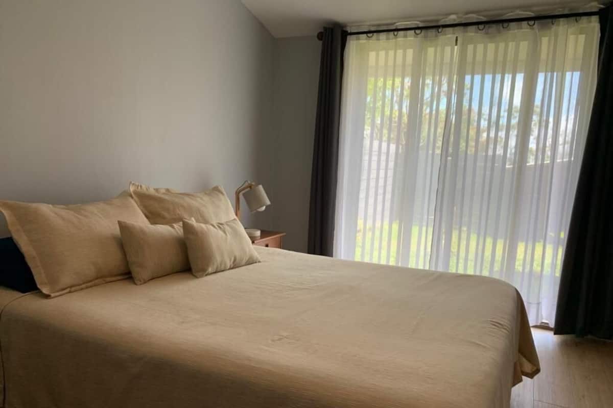 Un letto grande con copripiumino e cuscini color beige, un comodino con una lampada, e vista sul verde attraverso tende trasparenti.