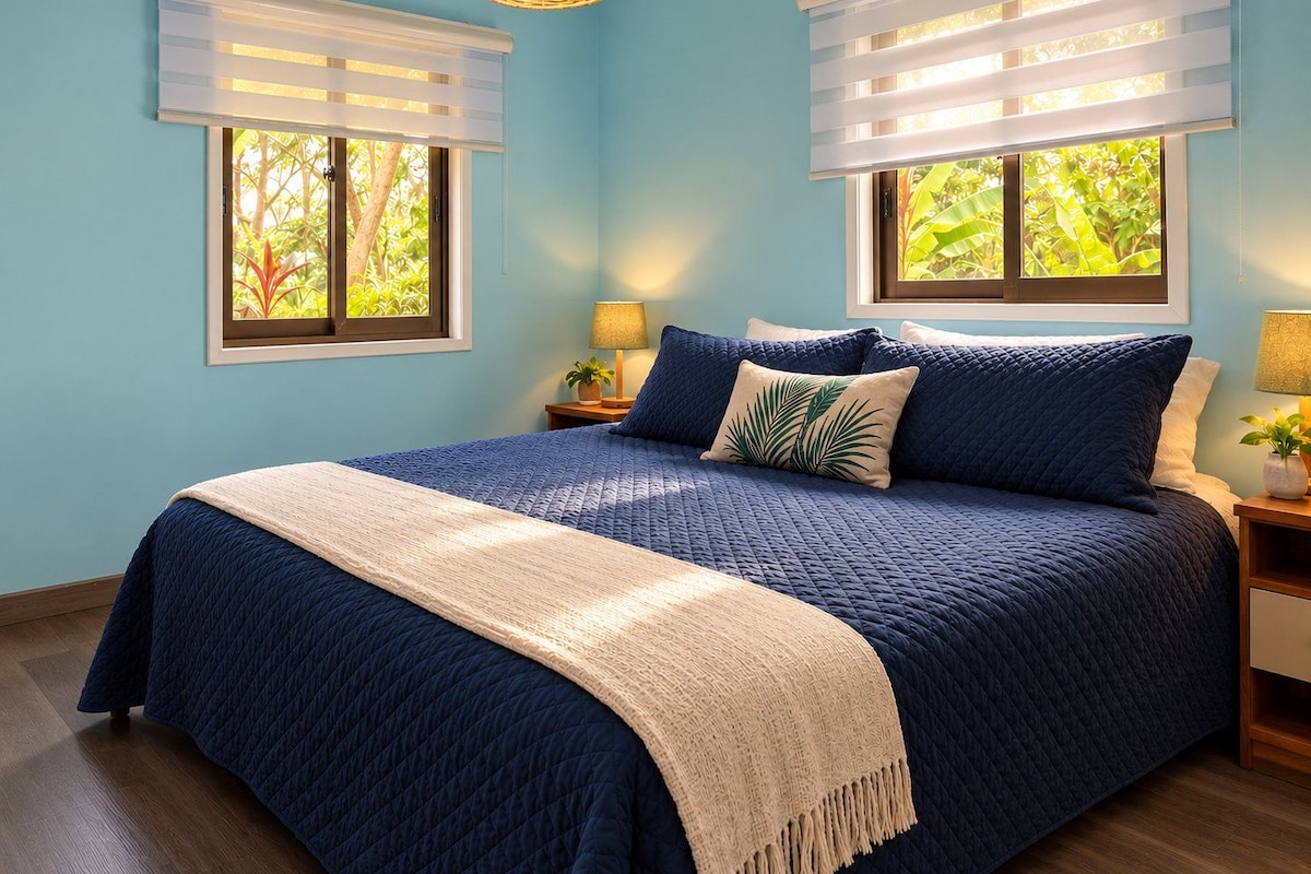 A bedroom with a large bed, two bedside tables with lamps, and two windows with blinds.