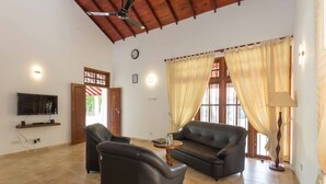 A spacious living room with a ceiling fan, a flat-screen TV mounted on the wall, a sofa, two armchairs, a coffee table, and a lamp. - Araliya Cottage (Negombo)