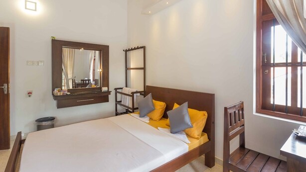 A bedroom with a bed, a mirror, a bedside table, and a window with curtains. - Araliya Cottage (Negombo)
