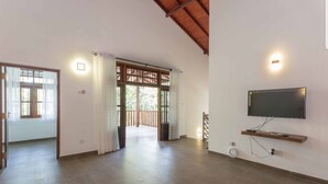 A spacious room with a wooden floor, a TV mounted on the wall, and a sliding door leading to an outdoor area. - Araliya Cottage (Negombo)