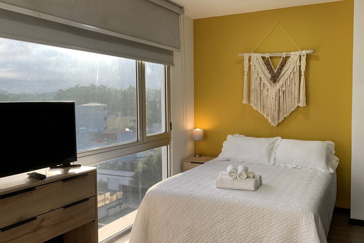 Ein Hotelzimmer mit einem Bett, einem Fernseher auf einer Holzkommode, einer Wanddekoration aus Makramee und Blick auf die Stadt durch große Fenster.