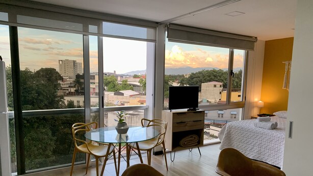 Ein modernes Hotelzimmer mit großem Balkon, einem Essbereich mit Glastisch und einem Bett mit weißer Decke.