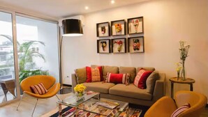 Ein modernes Wohnzimmer mit einer Couch, zwei Sesseln, einem Couchtisch und einer Stehlampe. An den Wänden befinden sich gerahmte Kunstwerke und auf einem Beistelltisch steht eine Pflanze.