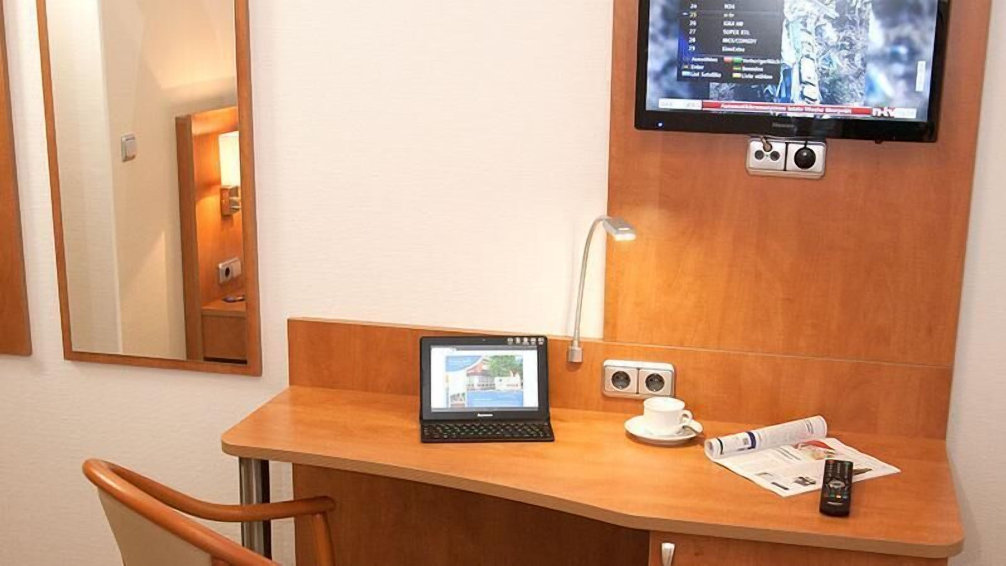 Habitación de hotel con escritorio de madera, una computadora portátil, televisor montado en la pared, mesita de noche con taza y control remoto, y un espejo.