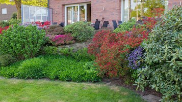 Un jardín bien cuidado con diversas plantas y arbustos florecientes frente a un edificio de ladrillo con puerta de cristal.