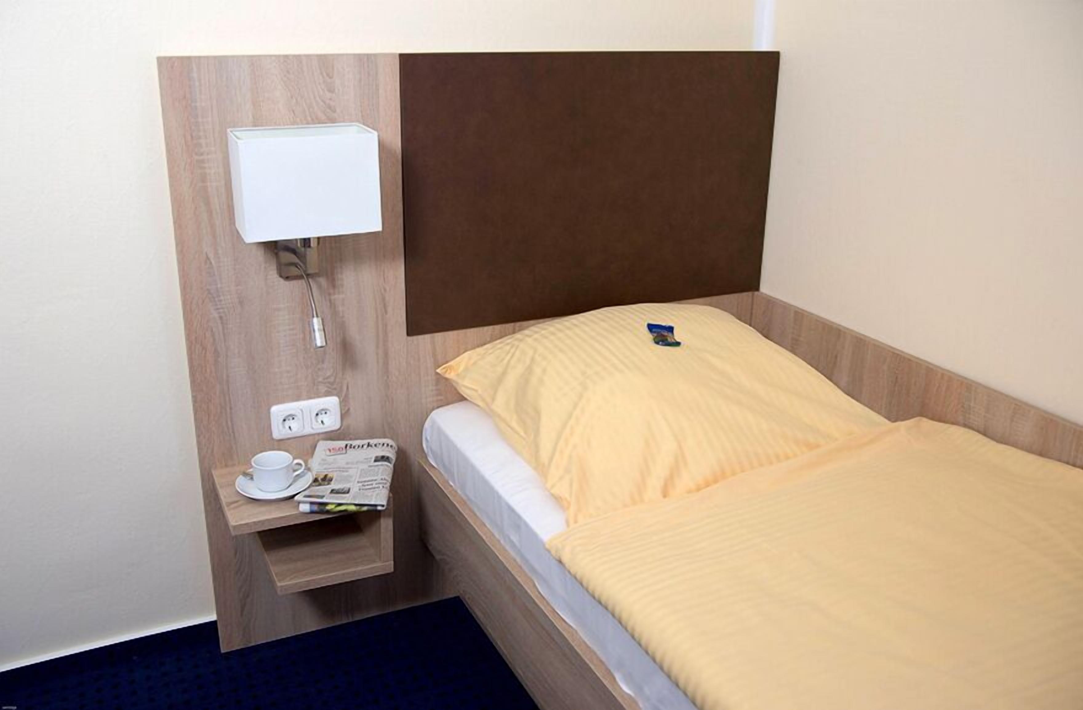 Hotellihuone, jossa on sänky, yöpöytä, jossa on kuppi ja sanomalehti, sekä seinään kiinnitetty valaisin.