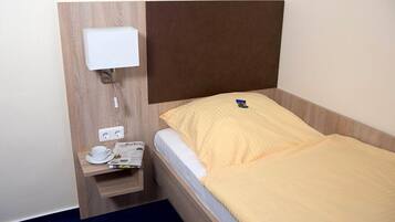 Hotellihuone, jossa on sänky, yöpöytä, jossa on kuppi ja sanomalehti, sekä seinään kiinnitetty valaisin.