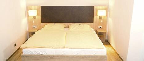 Una cama doble con estructura y cabecera de madera, dos mesitas de noche con lámparas y una pared de tono claro.