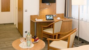 Hotellihuone, jossa on puinen työpöytä, kannettava tietokone, tuoli, pieni pöytä, jonka päällä on pullo ja hedelmiä, sekä seinälle asennettu televisio.