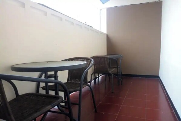 Un balcón pequeño y cerrado con dos mesas redondas y cuatro sillas de mimbre. El piso está cubierto de baldosas rojas y las paredes son lisas.