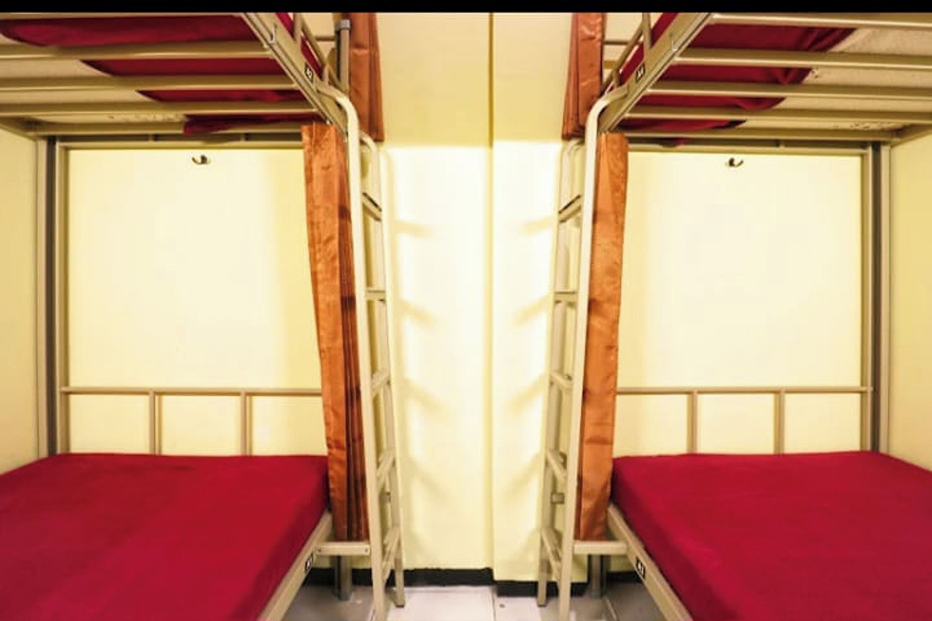 A train compartment with two single beds, each with a red bedspread, metal frames, and curtains.