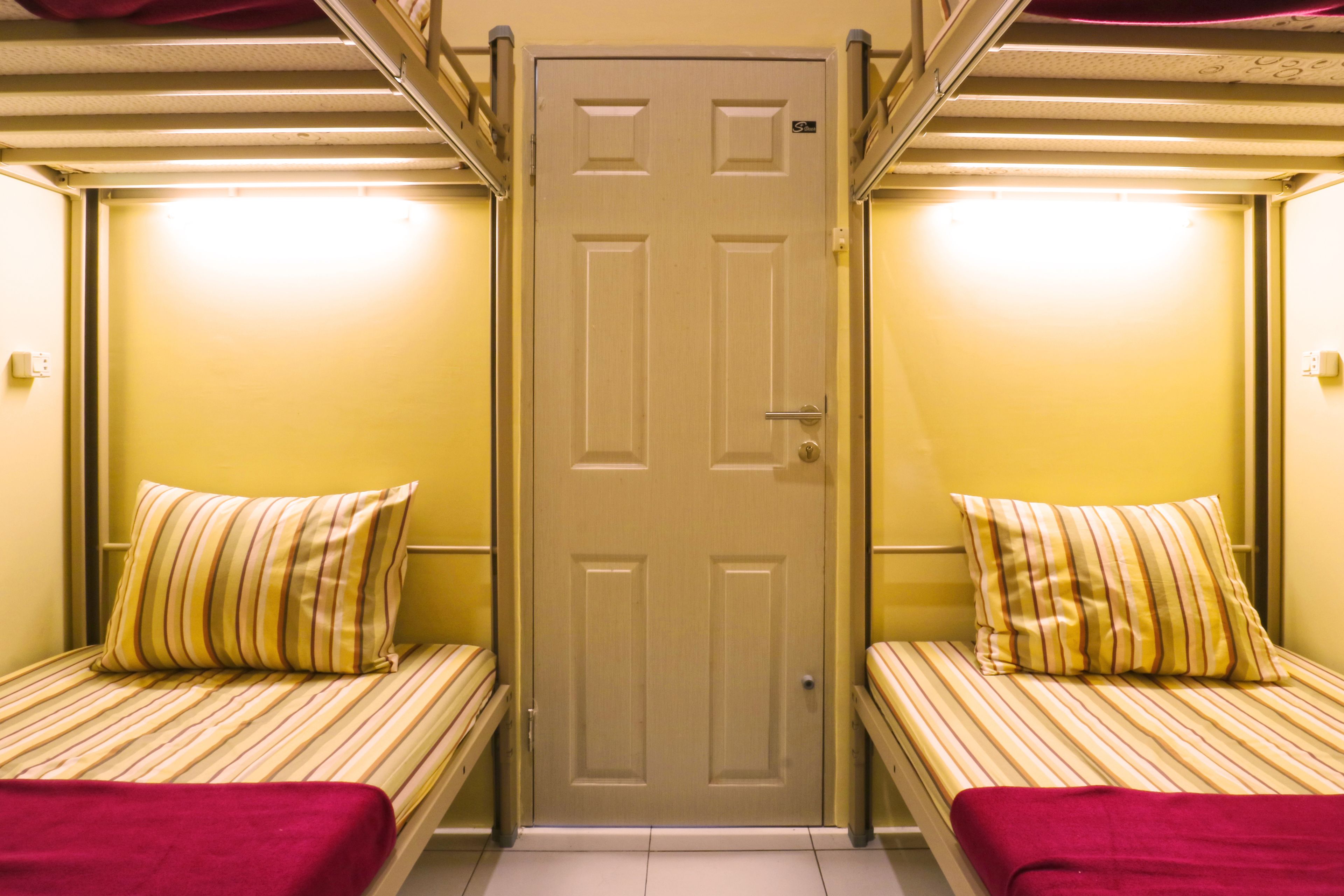 Una habitación con dos camas, cada una con ropa de cama a rayas y una manta roja. Una puerta con picaporte y cerradura está centrada entre las camas.