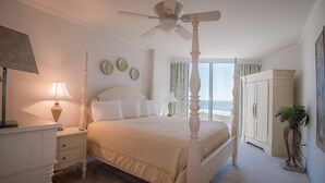 Uma cama com dossel, um abajur sobre a mesa de cabeceira e vista para o oceano através de uma janela com cortinas.
