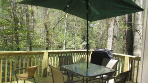 Relax on the deck with a clear view of the surrounding forest, featuring a glass-top table and four chairs, a grill, and a shaded area.