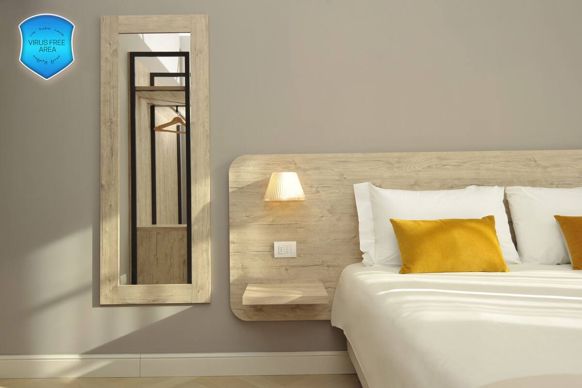 Una camera d'albergo con un letto, una lampada da comodino, uno specchio e un cartello con la scritta 'VIRUS FREE AREA'.