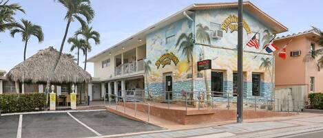 A building with a mural of palm trees and the ocean, a thatched-roof structure, and a sign indicating vacancy.