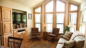 A cozy living room with a sofa, two armchairs, a coffee table, and a television. There are large windows with blinds, a wooden cabinet, and a rug on the floor. - Lillian Farms Bed & Breakfast (Washington)