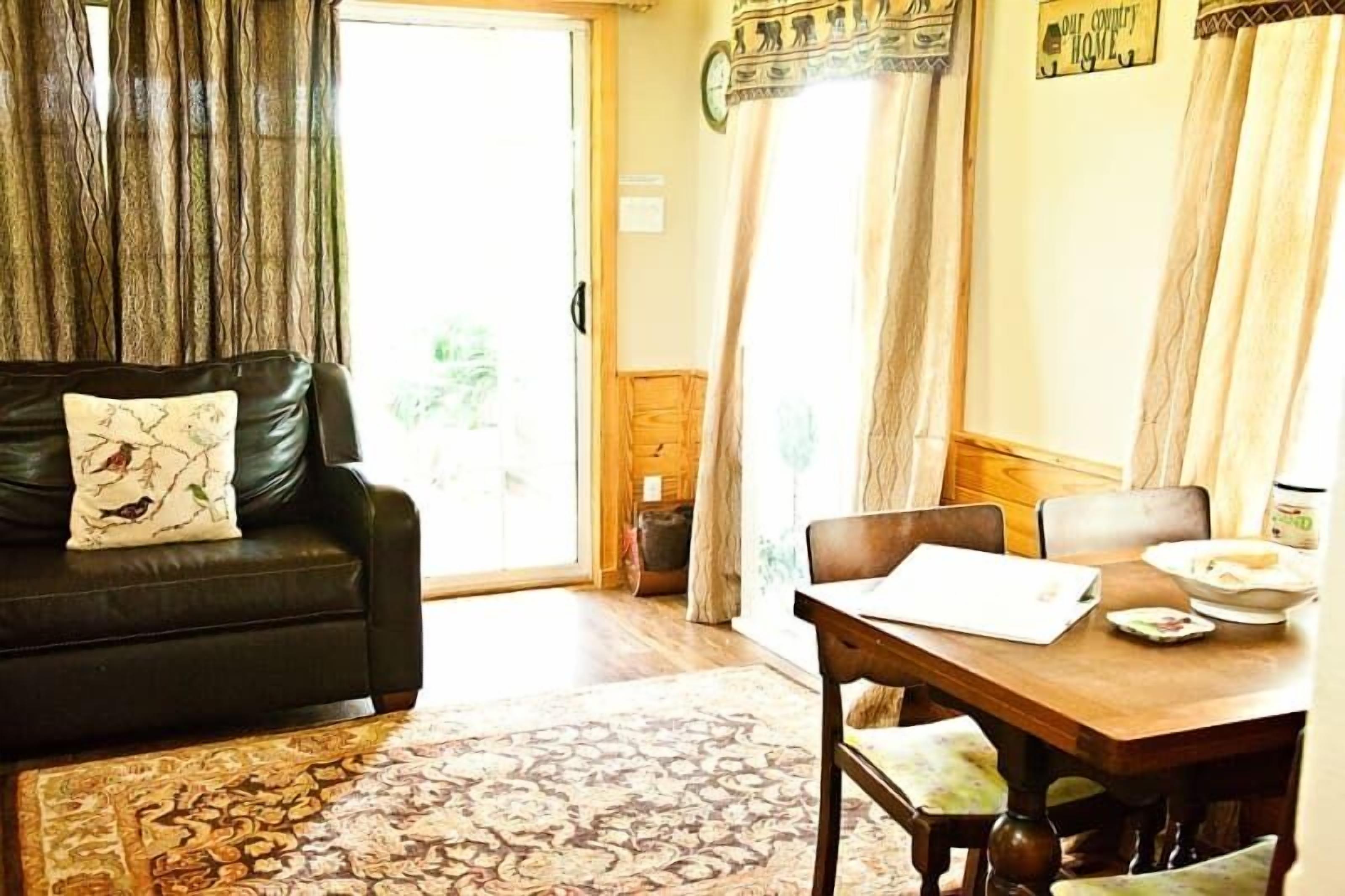 Sala de estar com sofá de couro preto, tapete estampado, mesa de jantar de madeira com cadeiras e vista por uma porta de vidro.
