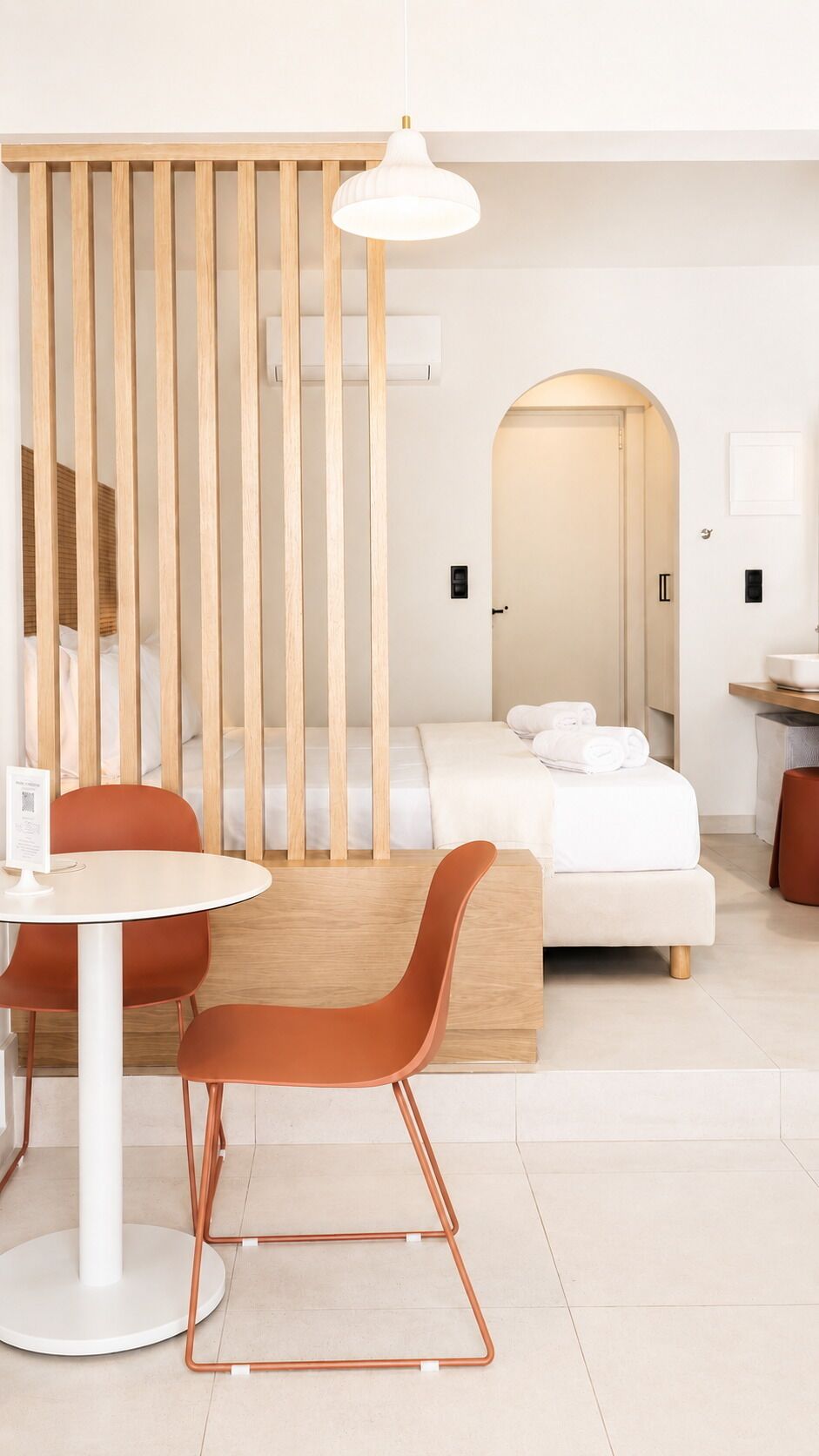 Une chambre d’hôtel moderne avec une cloison en bois, une table ronde blanche, deux chaises orange, un lit blanc et une lampe blanche.