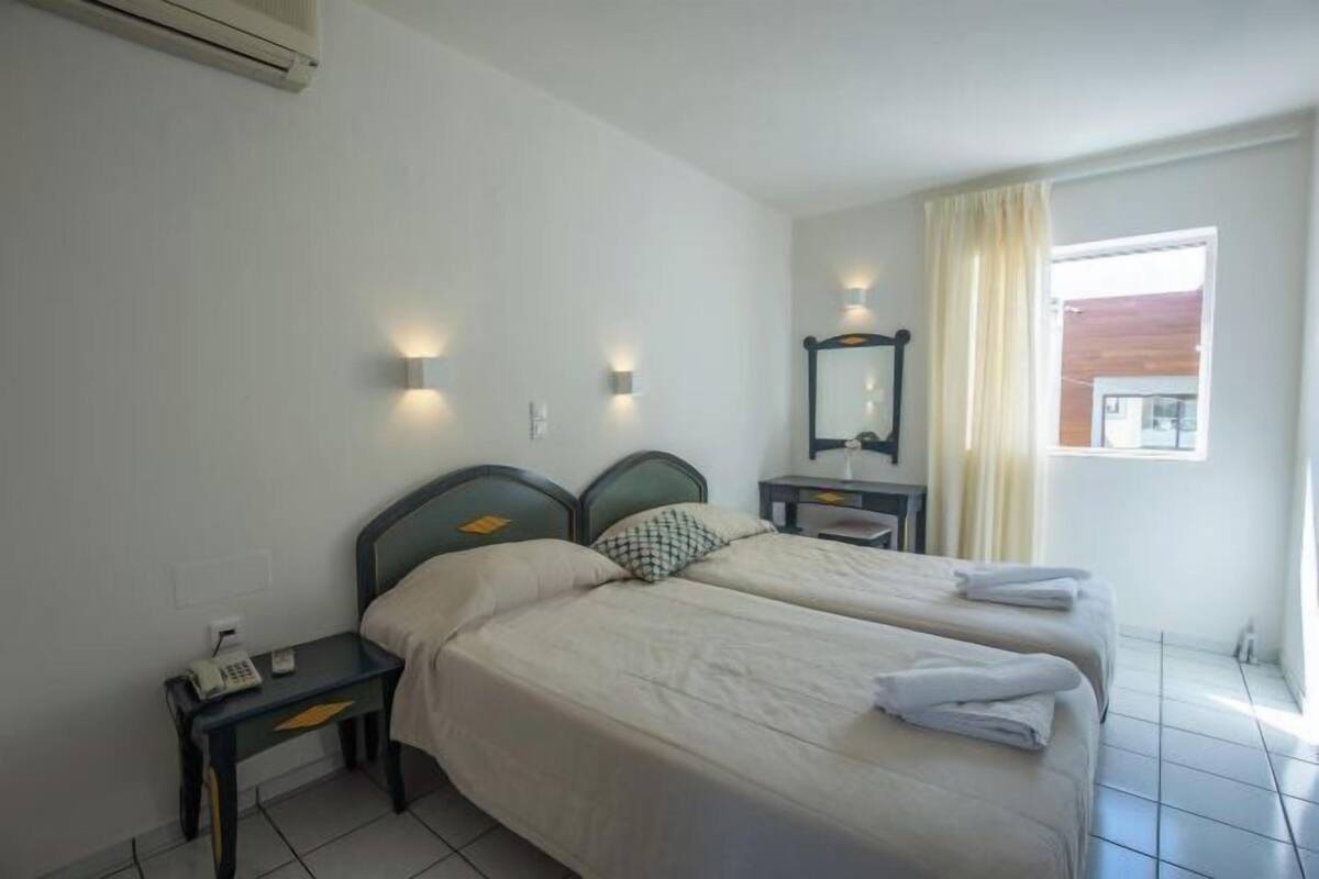 A hotel room with two beds, a bedside table with a phone, a mirror, and a window with curtains.