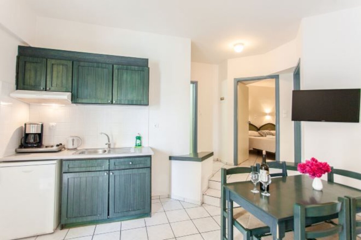 A compact kitchen with a sink, coffee maker, and cabinets. A dining table with chairs and a vase of flowers is visible.