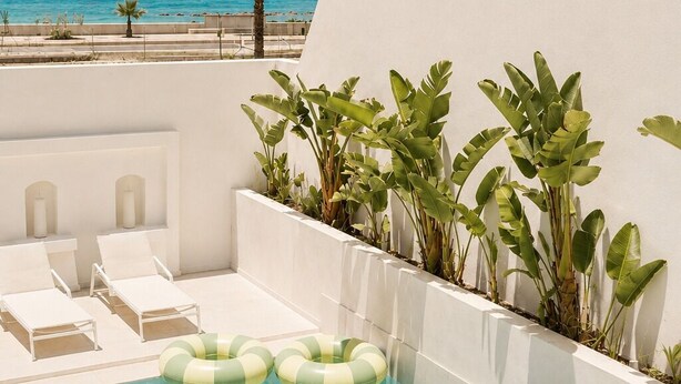 Un área de piscina con agua cristalina, sillas de descanso blancas y plantas tropicales.