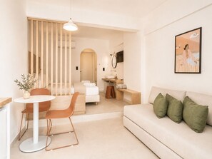 A modern hotel room with a bed, a sitting area with a table and chairs, a sofa, a small bathroom, and a painting on the wall. - Melitti Lucent (Rethymno)